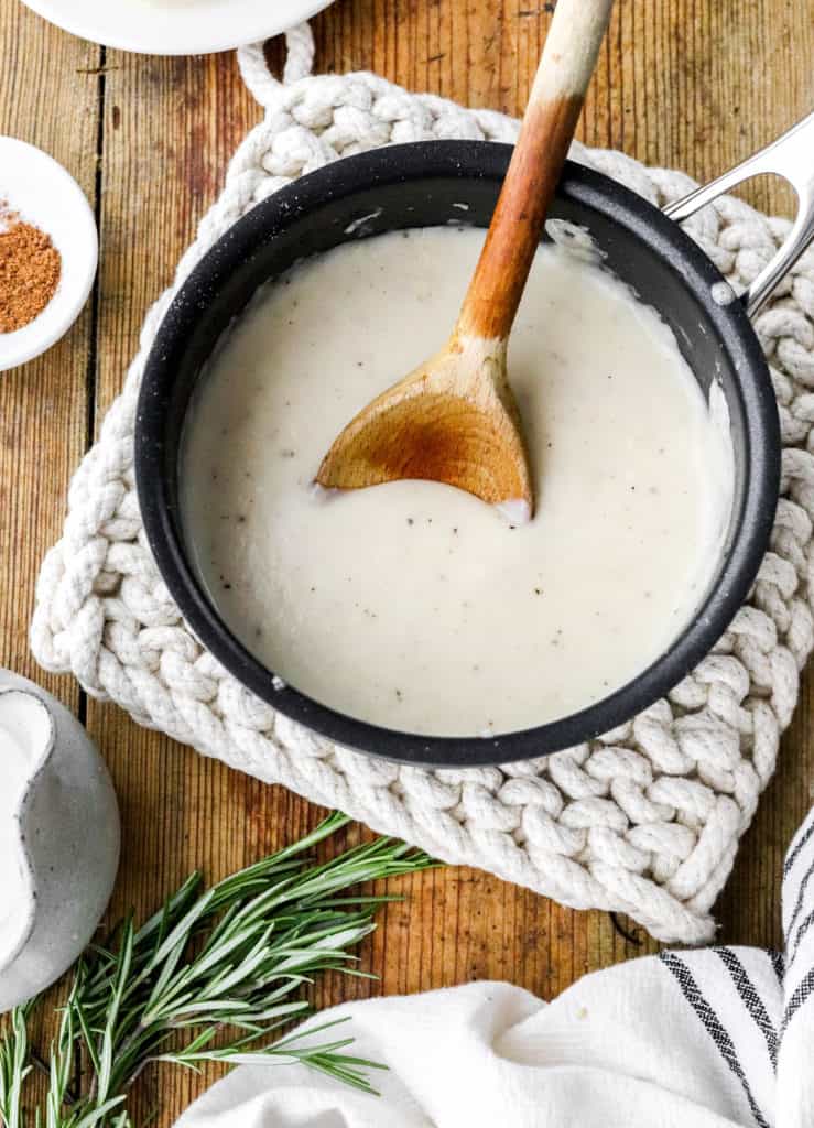 Dark sauce pan filled with cream sauce on top of a knitted white trivet with a bundle of herbs and some milk in a jar in front of it. 