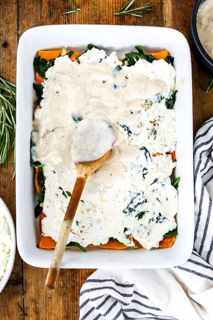 Wooden spoon spreading white cream sauce over lasagna in a white baking dish on a wooden board. 