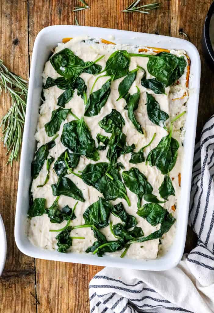 White rectangle baking dish filled with creamy white sauce with cooked spinach leaves spread out over the sauce with a striped linen and herbs next to the dish. 