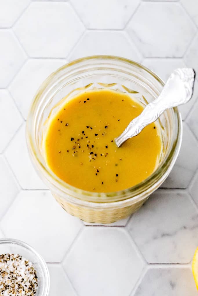 Glass mason jar filled with yellow dressing with a spoon in it on a marble surface