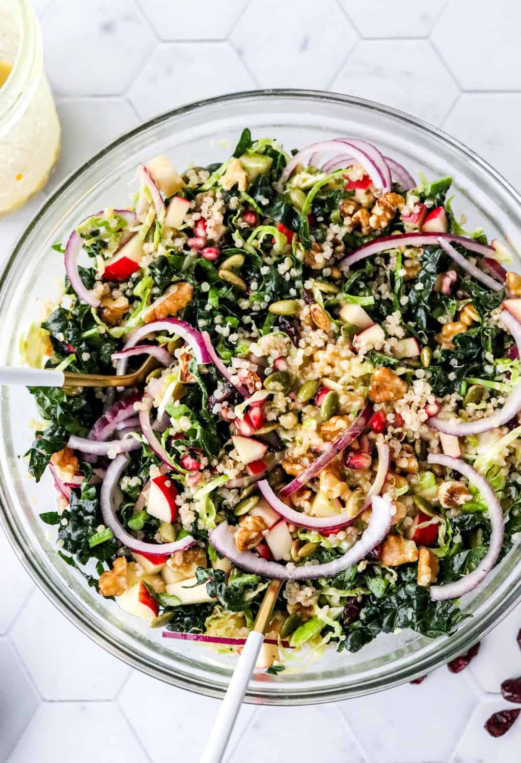 autumn kale salad with apple, sliced red onion, quinoa, nuts and seeds in a glass salad bowl with white and gold spoons in it on a marble countertop with a jar of dressing next to it. 