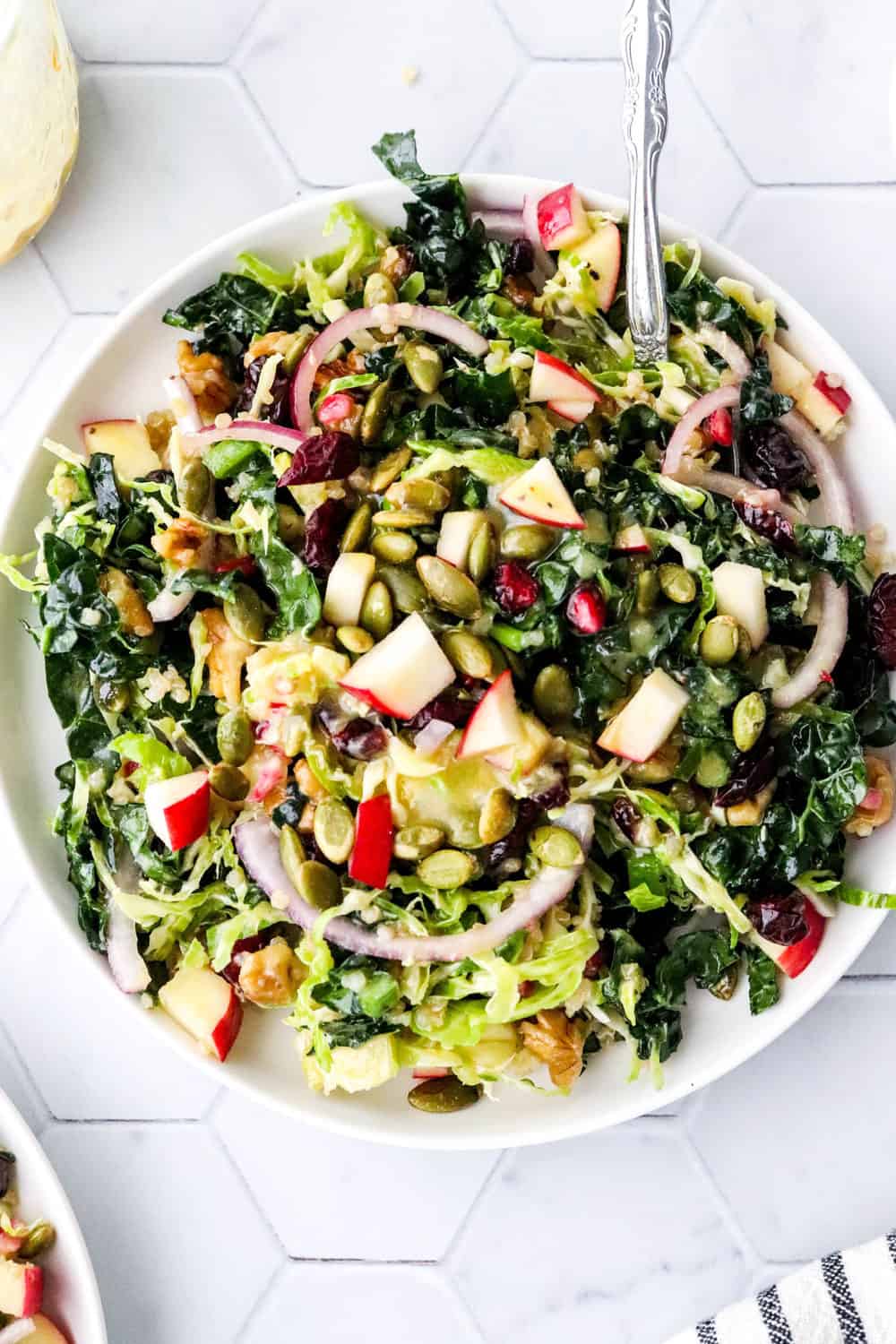 Round white plate with chopped kale, apple, onion pumpkin seeds and Brussel sprouts on it drizzled with mustard dressing with a silver fork on the plate. 
