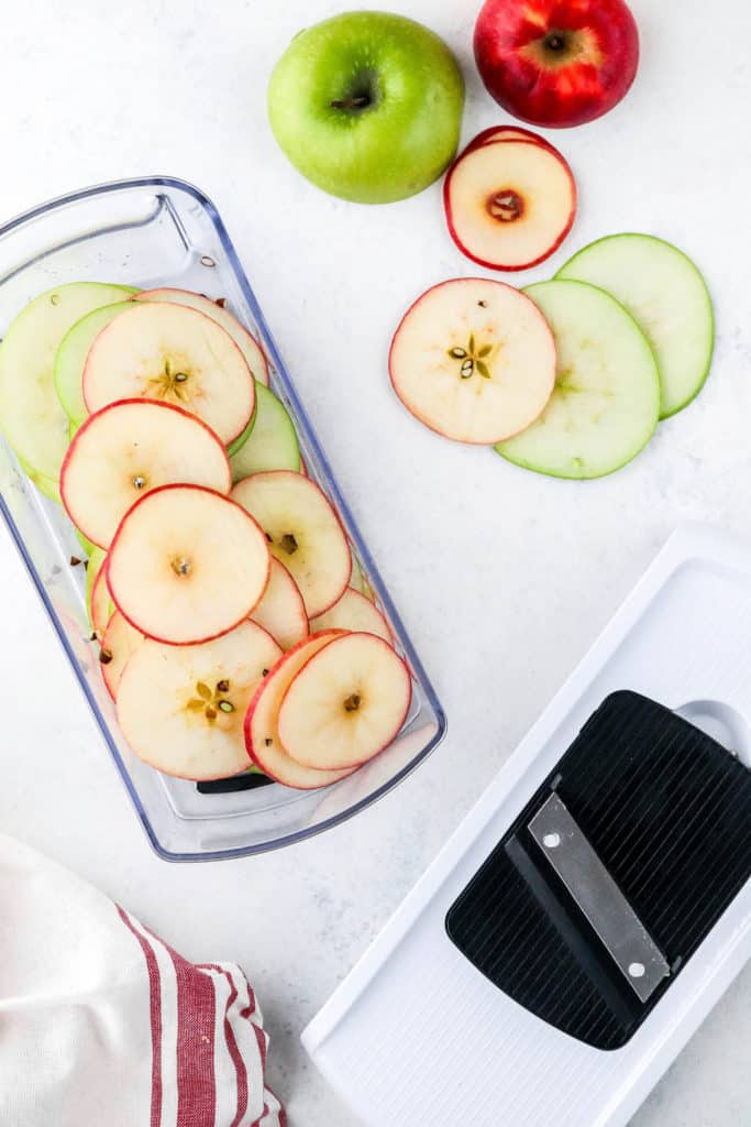 Long narrow plastic container filled with sliced green and red apples with more sliced apples next to it and a mandolin slicer in front of it. 
