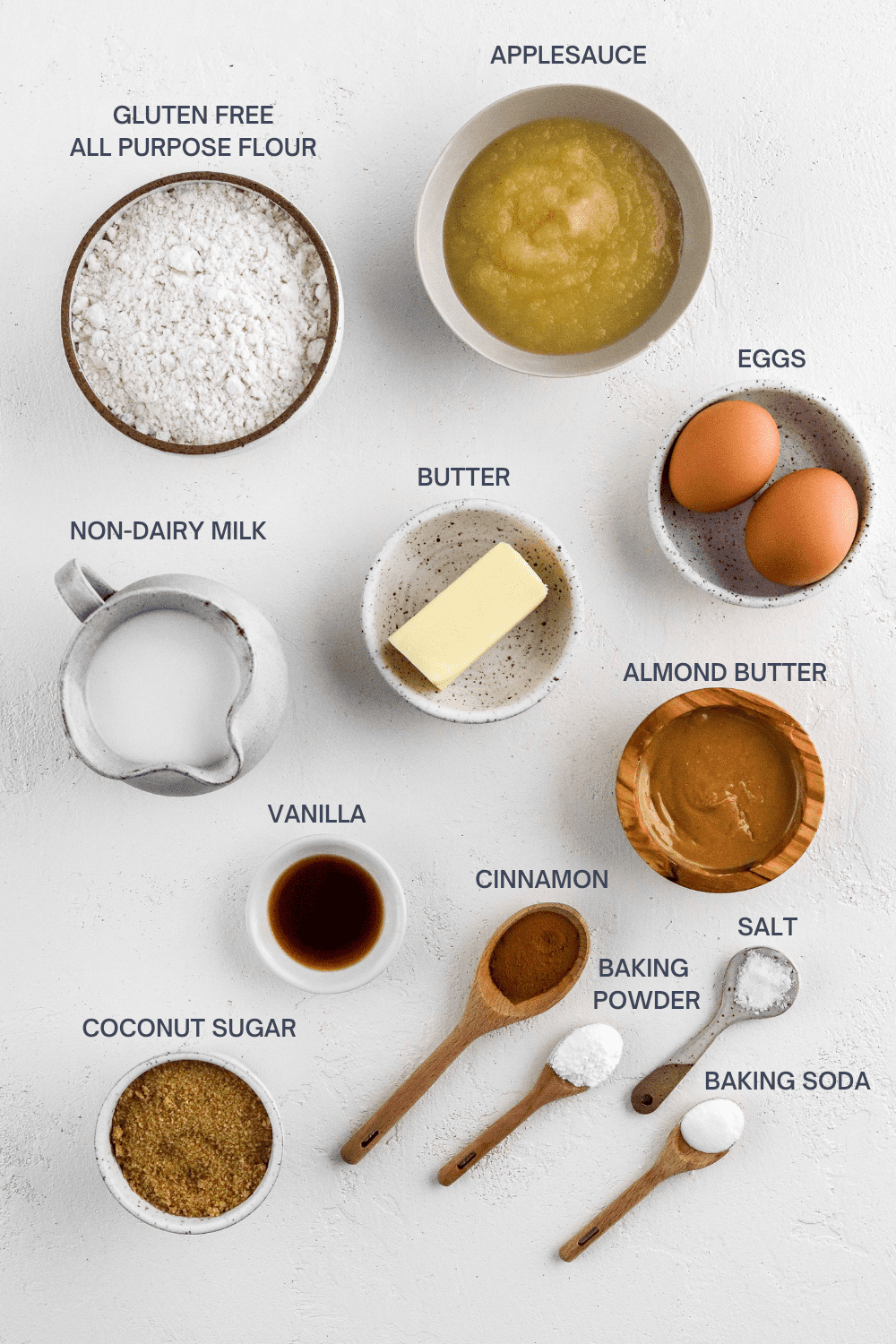 Bowl of flour next to a bowl of applesauce with a bowl with 2 eggs, plate with a stick of butter, cup of milk, brown bowl with almond butter, small bowl of vanilla and spoons with cinnamon, salt, baking powder and baking soda in them with labels for each ingredient all on a white surface. 