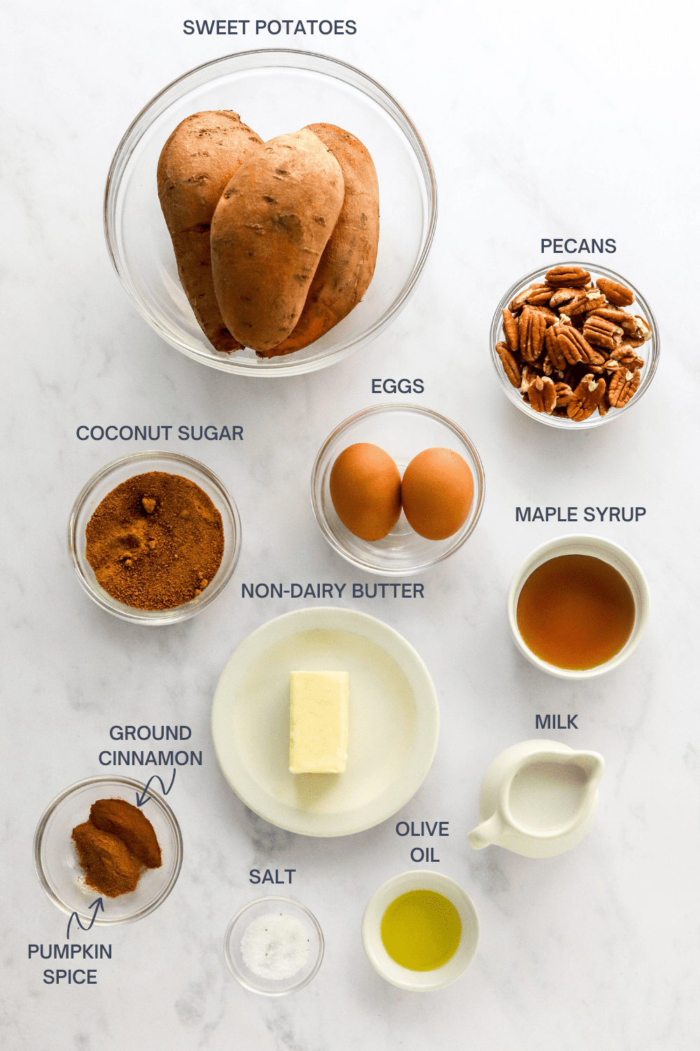 Bowl of whole sweet potatoes, bowl of pecans, bowl of coconut sugar, bowl of whole eggs, bowl with maple syrup and a plate with a stick butter on a white marble surface. 