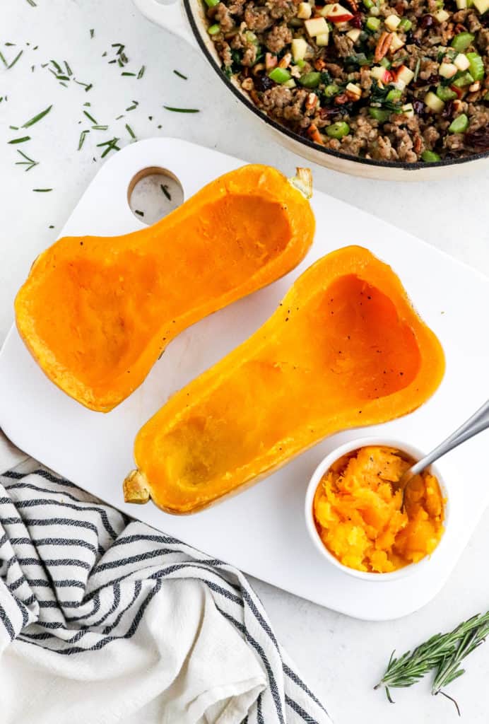 Two halves of cooked butternut squash with the center scooped out on a white cutting board with a striped towel in front of it and a round pan with sausage stuffing behind it