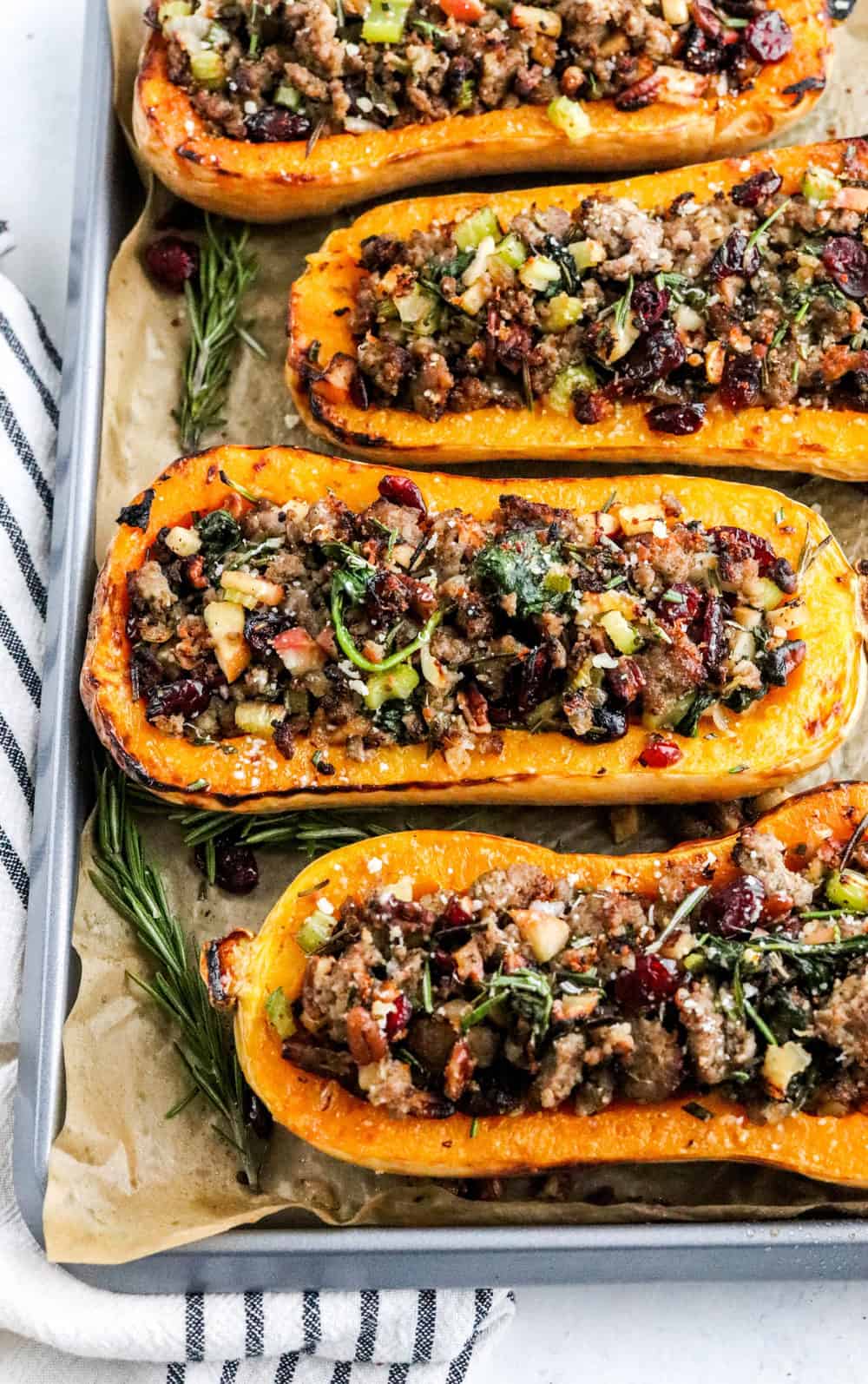 Halved squash filled with sausage stuffing on a baking sheet with large pieces of fresh rosemary next to is and a white and blue striped towel under the baking sheet. 