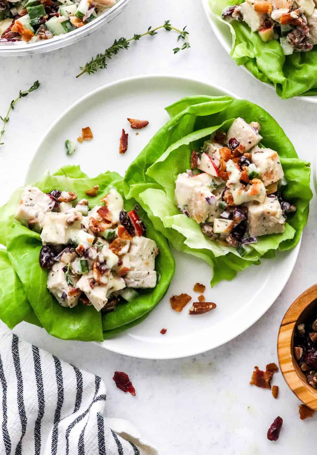 creamy cranberry chicken salad in green lettuce cups on a round white plate with another plate with more chicken salad behind it and a sprig of time on the table in back of it. 
