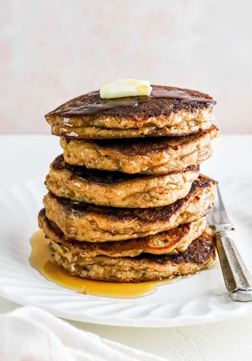 Stack of dark brown apple cinnamon pancakes covered in syrup with butter on top of them on a round white plate. 
