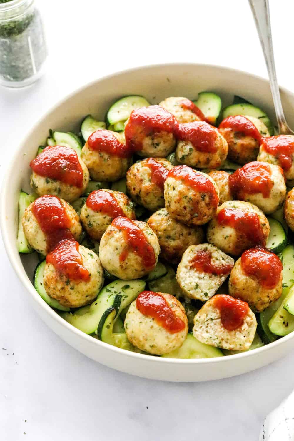 Round tan bowl filled with a pile of sliced, cooked zucchini with cooked golden brown meatballs on top of the zucchini drizzled with red sauce with a spoon in the dish. 
