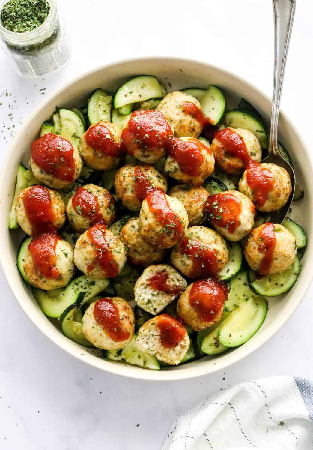 Round cream coloured dish filled with sliced zucchini topped with ground chicken meatballs slathered with red sauce with a spoon in the bowl and a glass jar of green herbs behind it. 