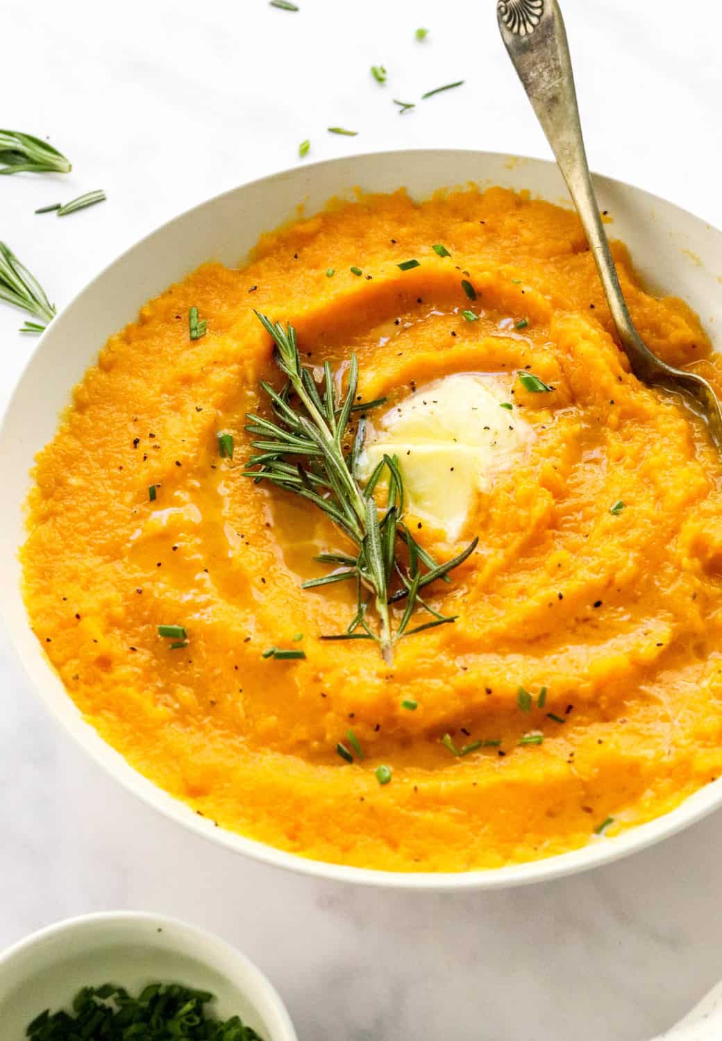 White serving bowl filled with orange mashed squash with butter and rosemary on top of it with a spoon in the bowl on a white surface.