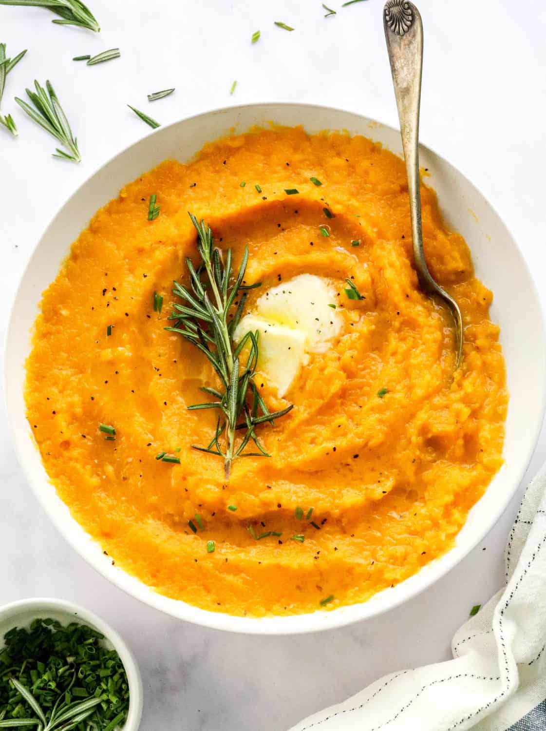 White bowl with mashed butternut squash garnished with some melty butter and herbs with a bowl of chives and a linen in front of it and some rosemary leaves scattered behind it. 