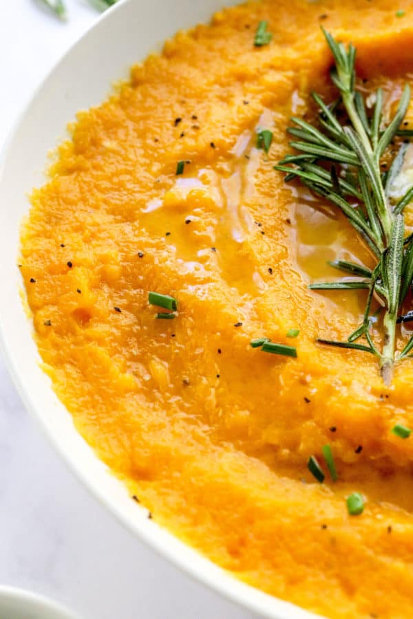 Close up shot of fluffy mashed squash in a white bowl topped with a sprig of fresh rosemary