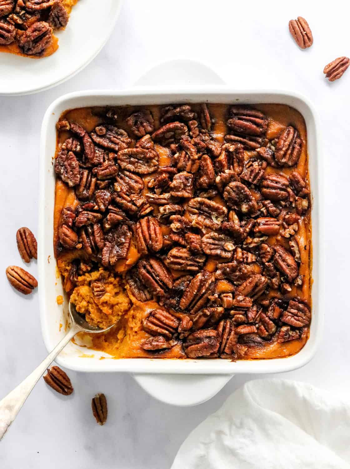 Healthy sweet potato casserole in a square, white dish topped with pecans with a spoon in the dish and a plate of the casserole behind it. 