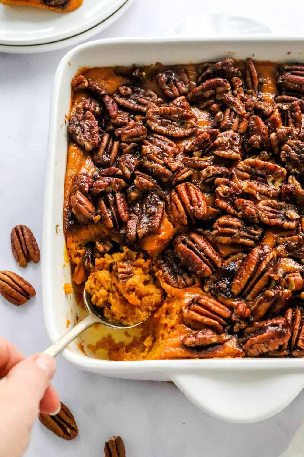 Hand using a spoon to scoop out some sweet potato casserole from a baking dish with white, stacked plates behind it and 2 pecans on the counter next to it. 