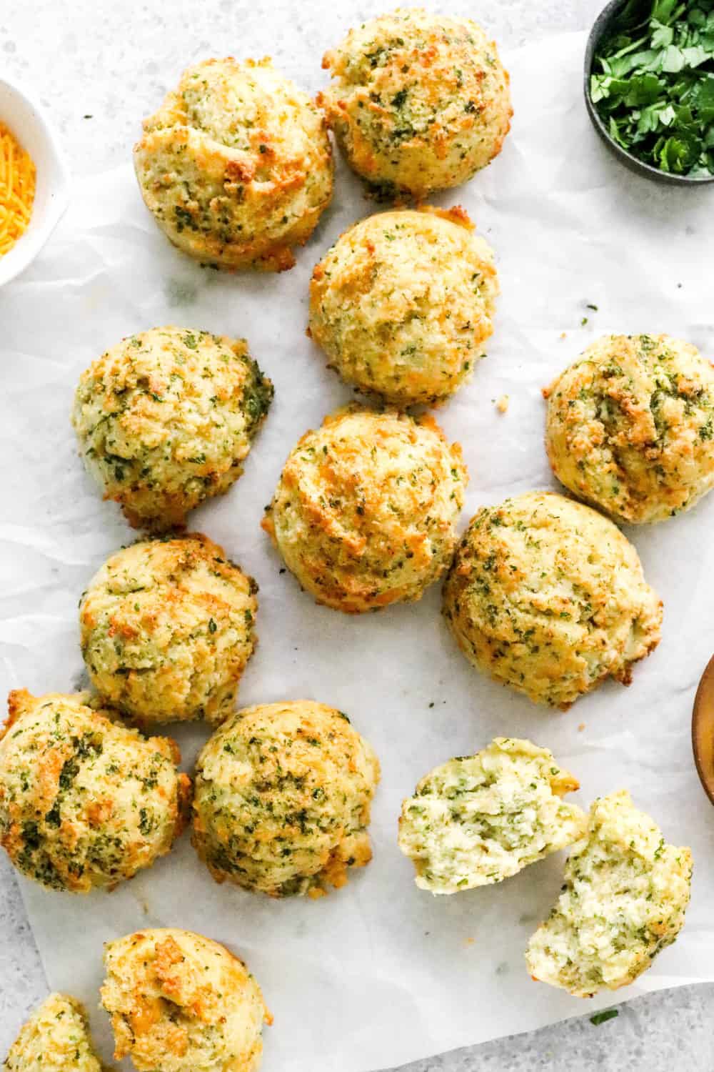 A bunch of homemade cheddar herb biscuits on white parchment paper with a bowl of shredded cheddar cheese and a bowl of parsley behind them. 