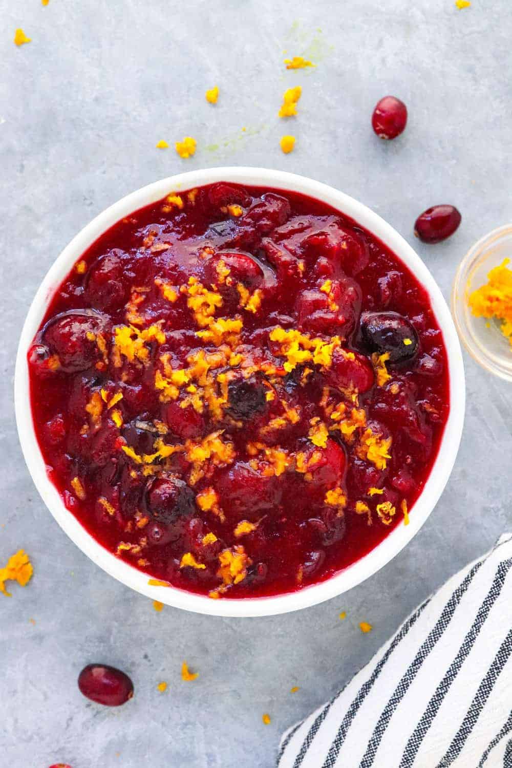 Round bowl filled with bright red cranberry sauce topped with orange zest with a few cranberries and orange zest sprinkled around it. 