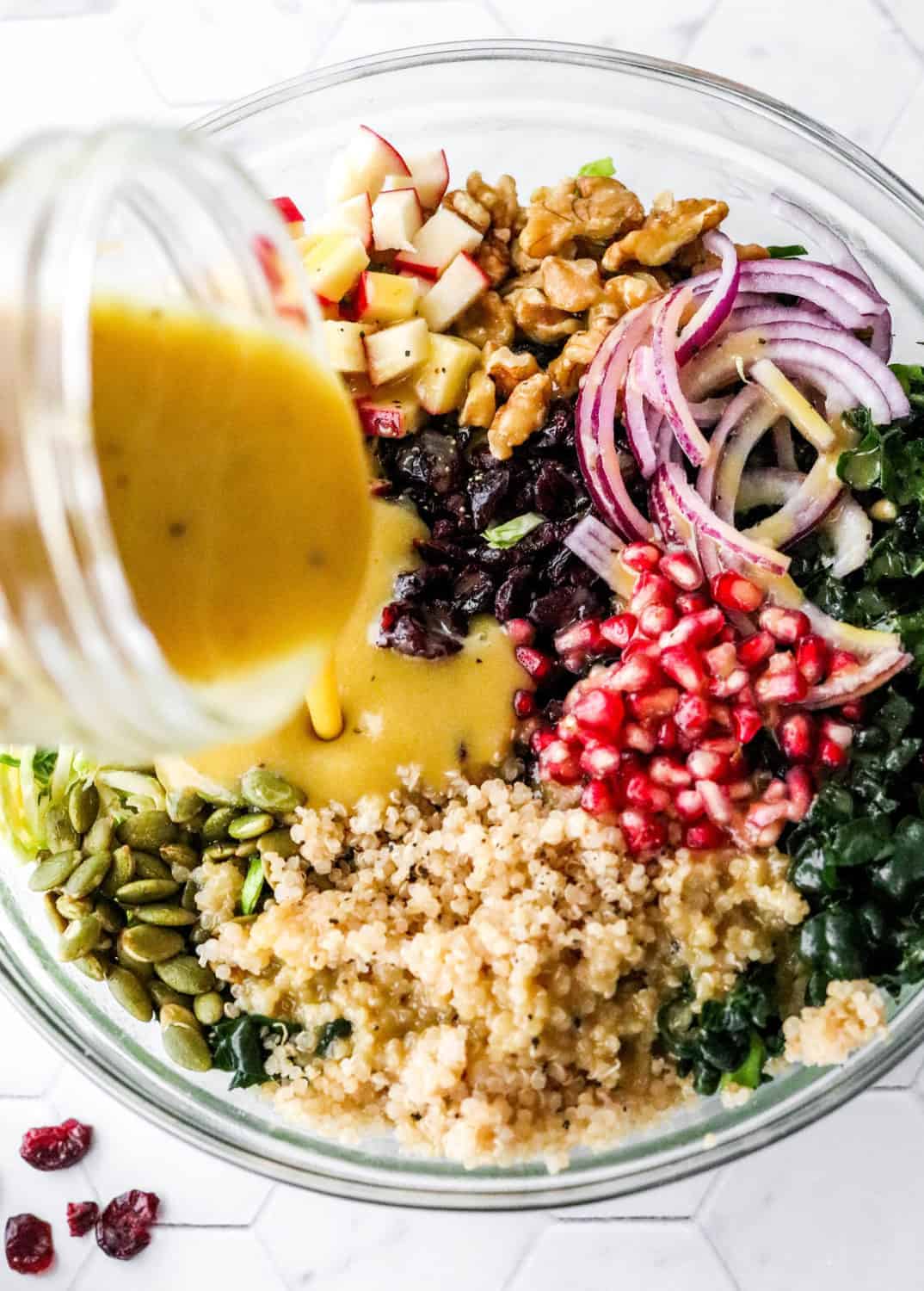 Jar pouring yellow dijon dressing over a bowl filled with kale, nuts, pomegranate seeds, cranberries and quinoa. 