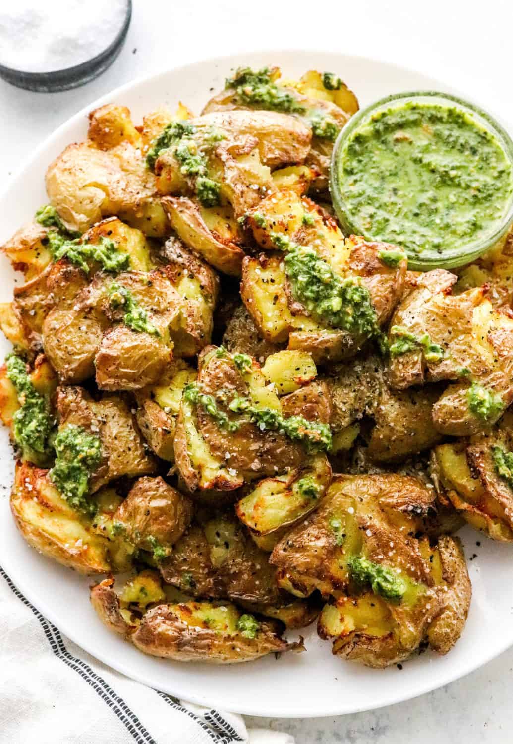 Air FRYER crispy smashed potatoes on a round platter and drizzled with pesto sauce with a bowl of salt behind it. 