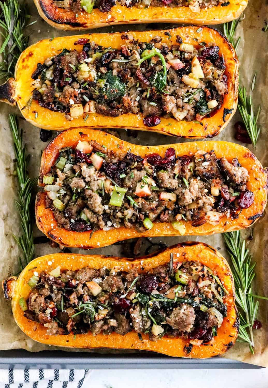 Three halves of roasted butternut squash filled with sausage apple filling on a baking sheet with fresh rosemary around it. 