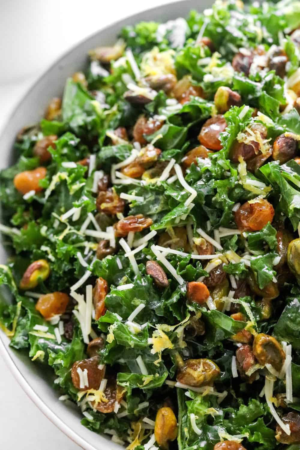 Kale salad in a serving bowl topped with raisins, nuts, lemon zest and parmesan cheese. 