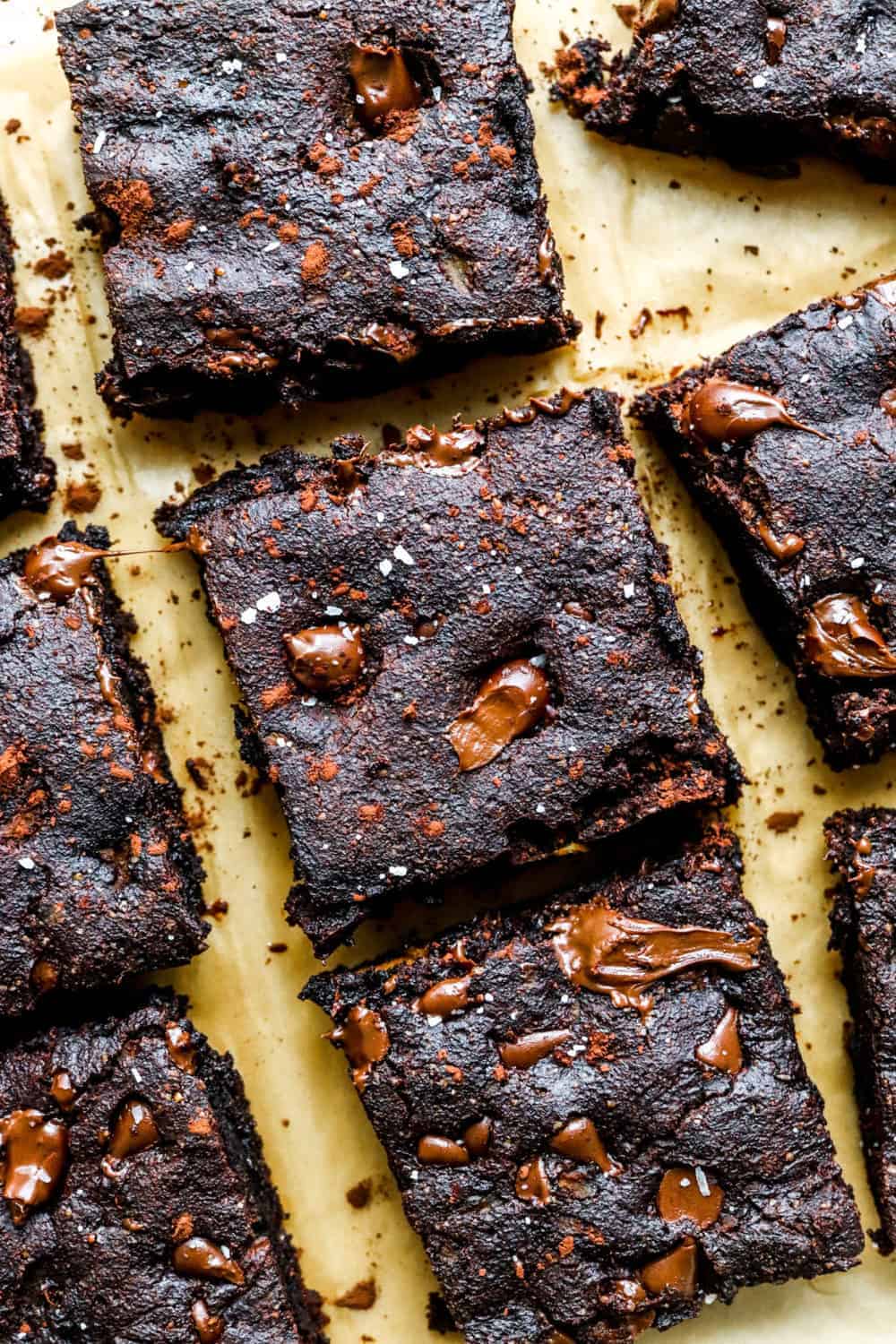 Dark chocolate sweet potato brownies in rows on brown paper with melty chocolate chips pressed into them. 