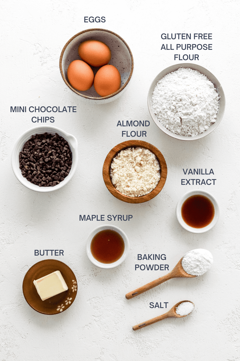 Three eggs in a brown, specked bowl, with a bowl of flour, bowl of mini chocolate chips, plate with a stick of butter, small bowl with maple syrup, bowl of vanilla, wooden spoon with baking powder and a small wooden spoon with salt in front of it. With labels of each ingredient above each item on a white surface. 