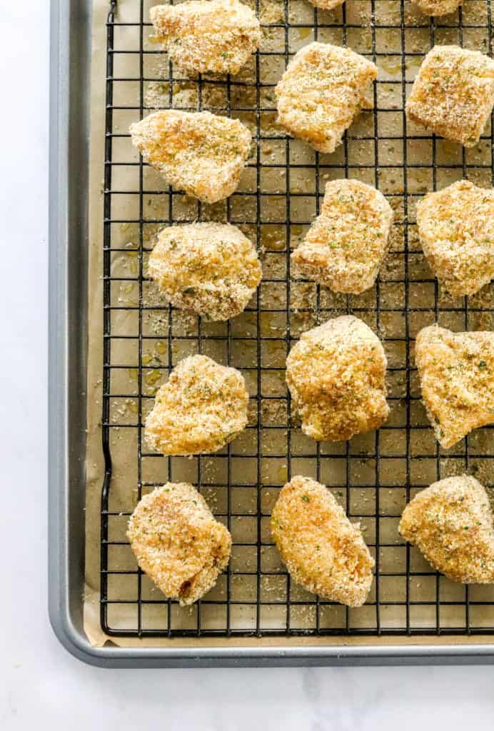 Breaded chicken nuggets on a black baking rack on top of a baking sheet lined with brown parchment paper. 
