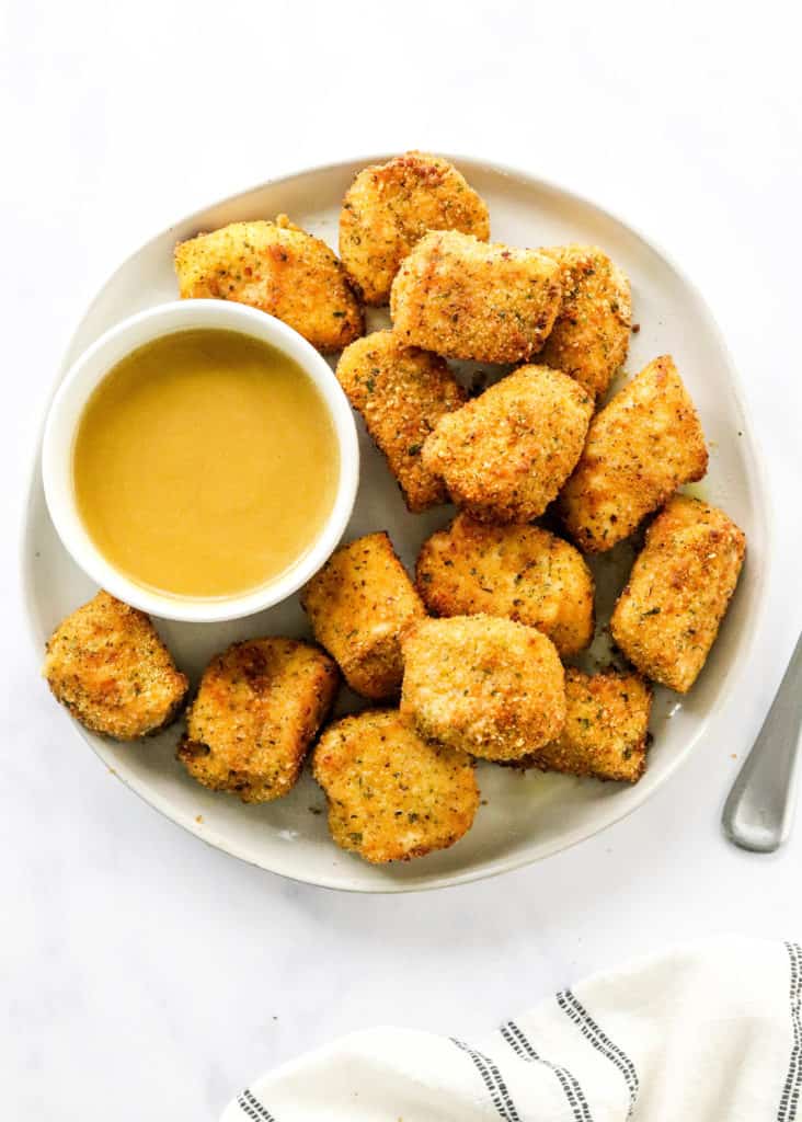 Round plate with cooked chicken nuggets on it next to a round white bowl filled with honey mustard sauce with a spoon next to the plate on a white surface. 