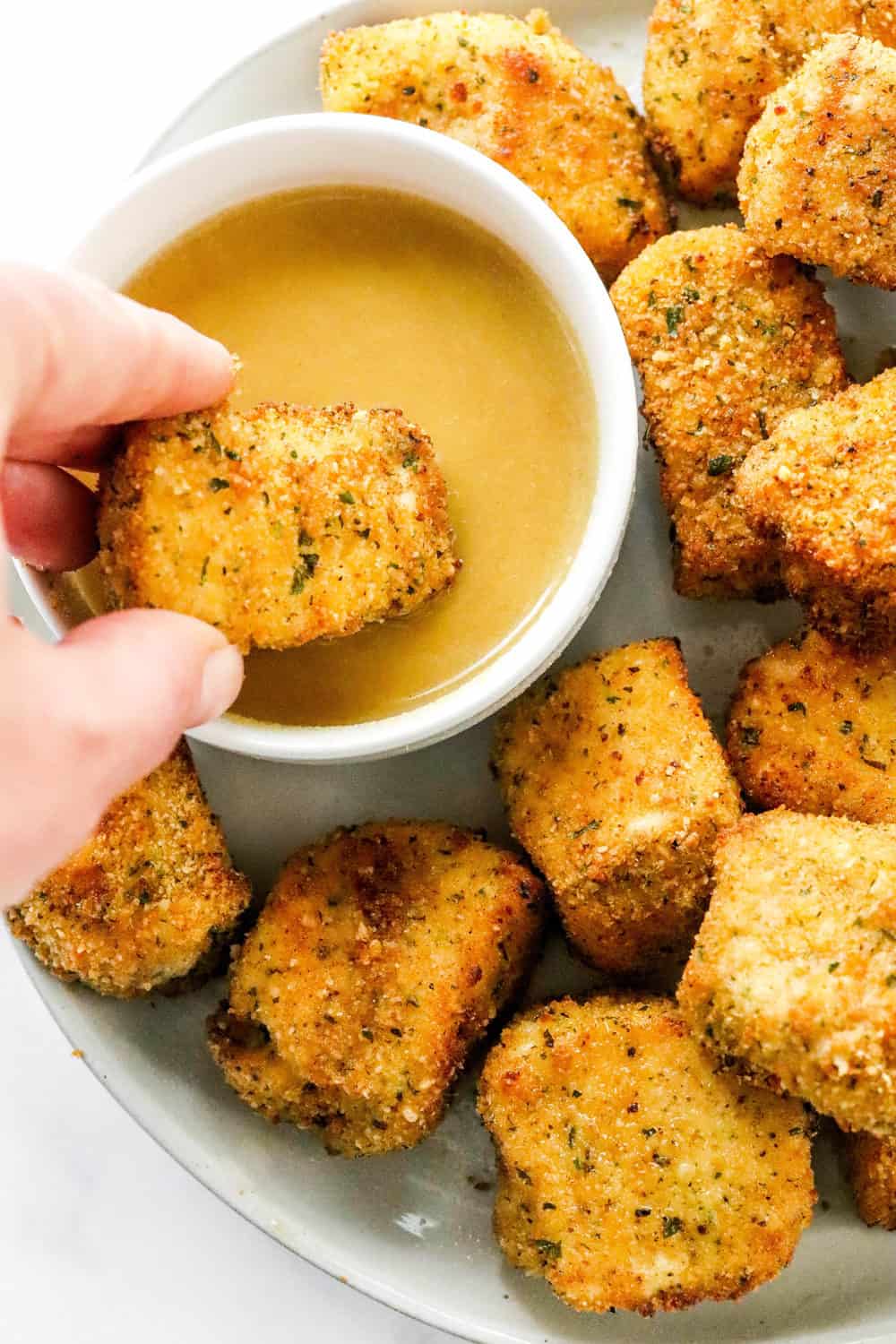 Hand dipping crispy chicken nuggets into a honey mustard sauce with more chicken nuggets on the plate. 