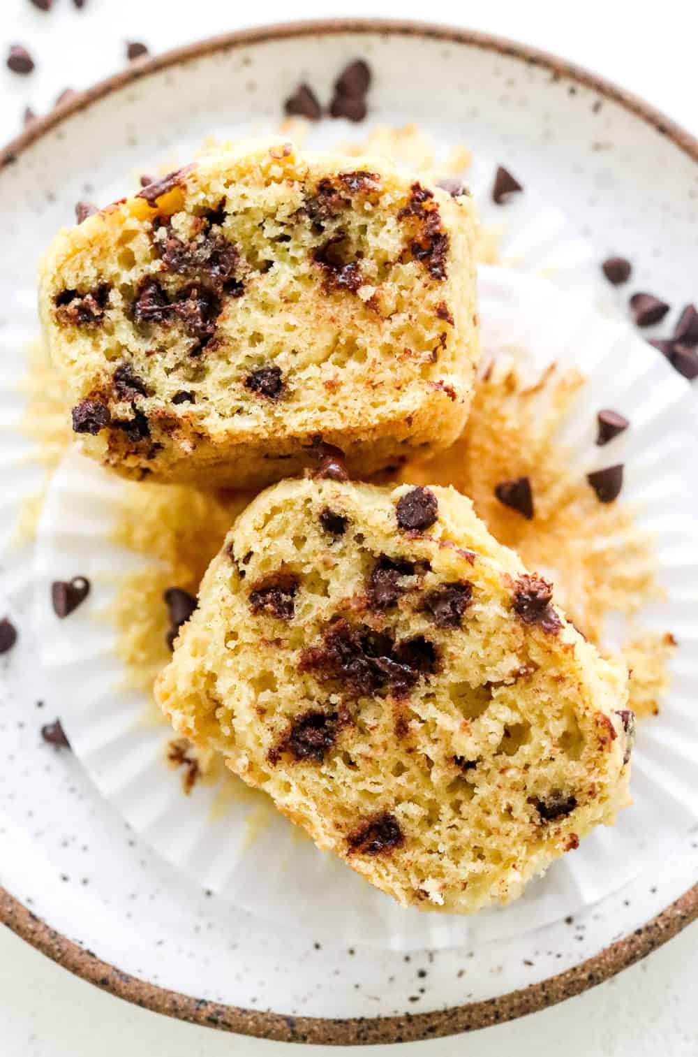 Chocolate chip muffin sliced open on a brown specked plate with chocolate chips sprinkled on the plate.
