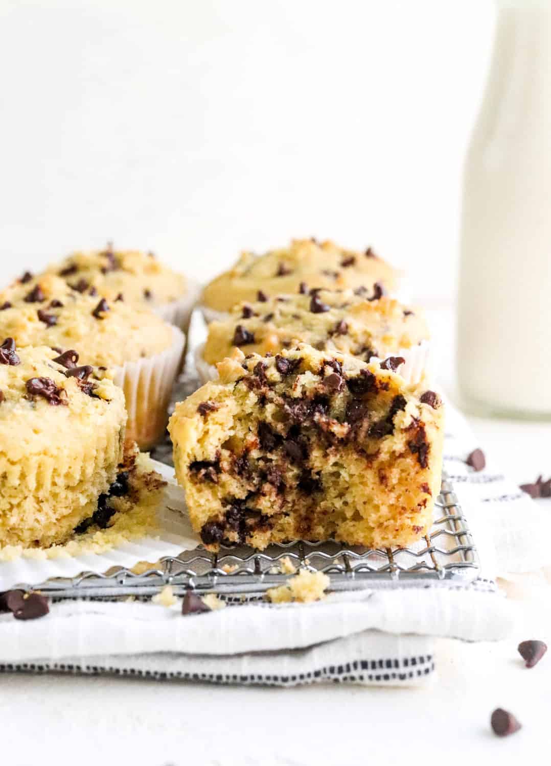 Tow rows of baked chocolate chip muffins with a bite taken out of one in the front on a wire rack on top of a white linen with more chocolate chips around it and a jug of milk behind it. 