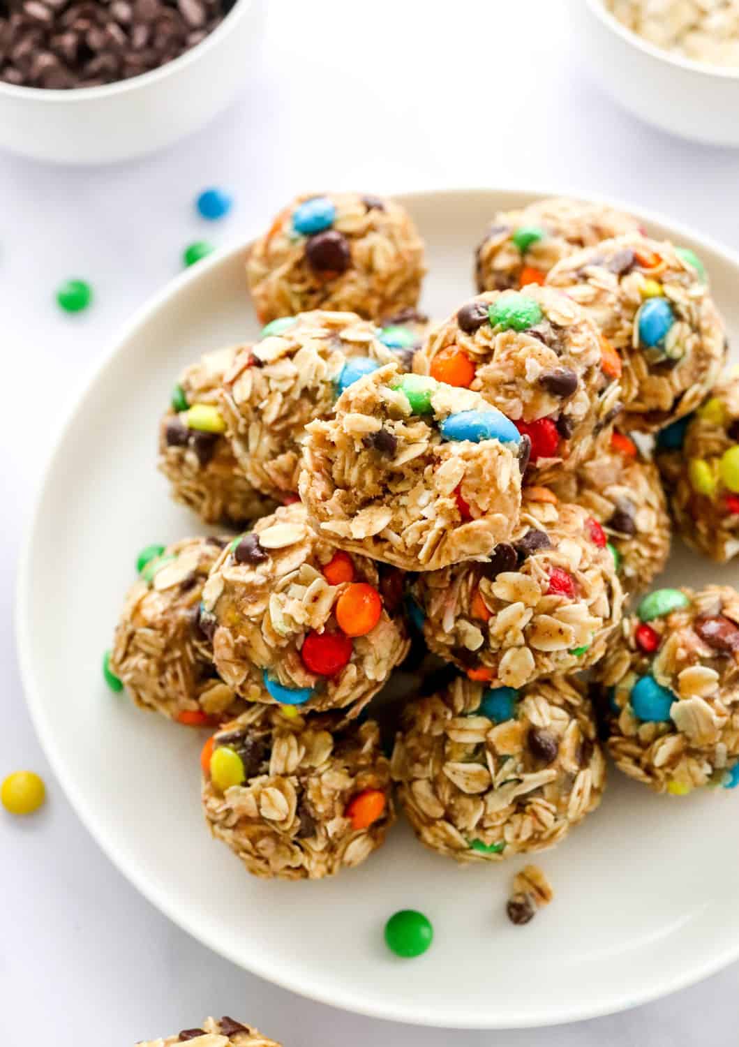 Monster cookie energy balls piled on top of one another on a round, white plate with a bowl if mini chocolate chips and a bowl of oats behind it and some mini m&ms scattered around it. 