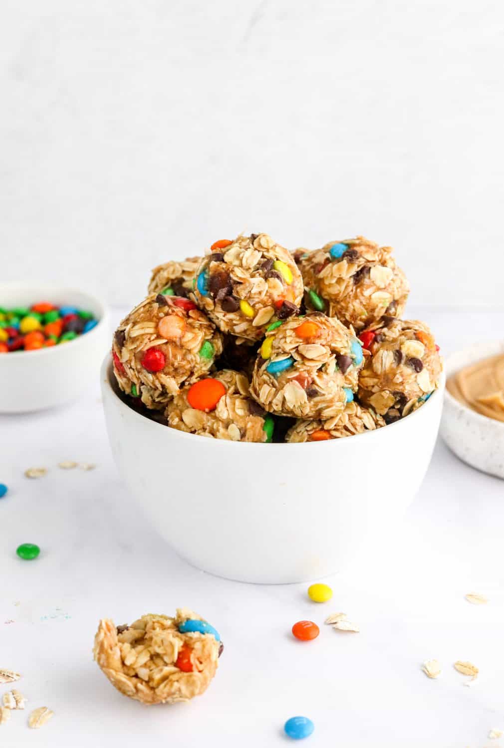 Healthy energy bites with m&ms in them in a white, round bowl with more  m&ms in a bowl behind it with a piece of an energy ball in front of the bowl. 