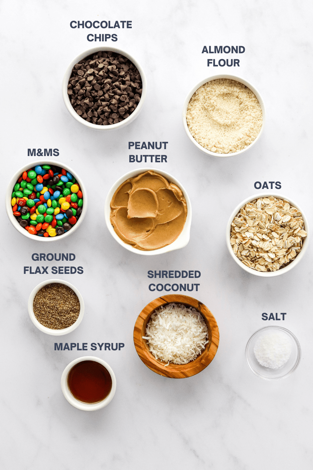 Round white bowl of mini chocolate chips with a bowl of almond flour, bowl of m&ms, bowl of peanut butter, bowl of oats, flax seeds, coconut, syrup and salt in front of it on a marble surface. 