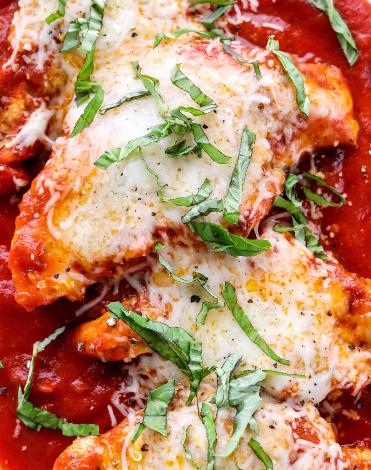 Pieces of saucey, cheese chicken on top of a best of tomato sauce with chopped basil on top of the chicken. 