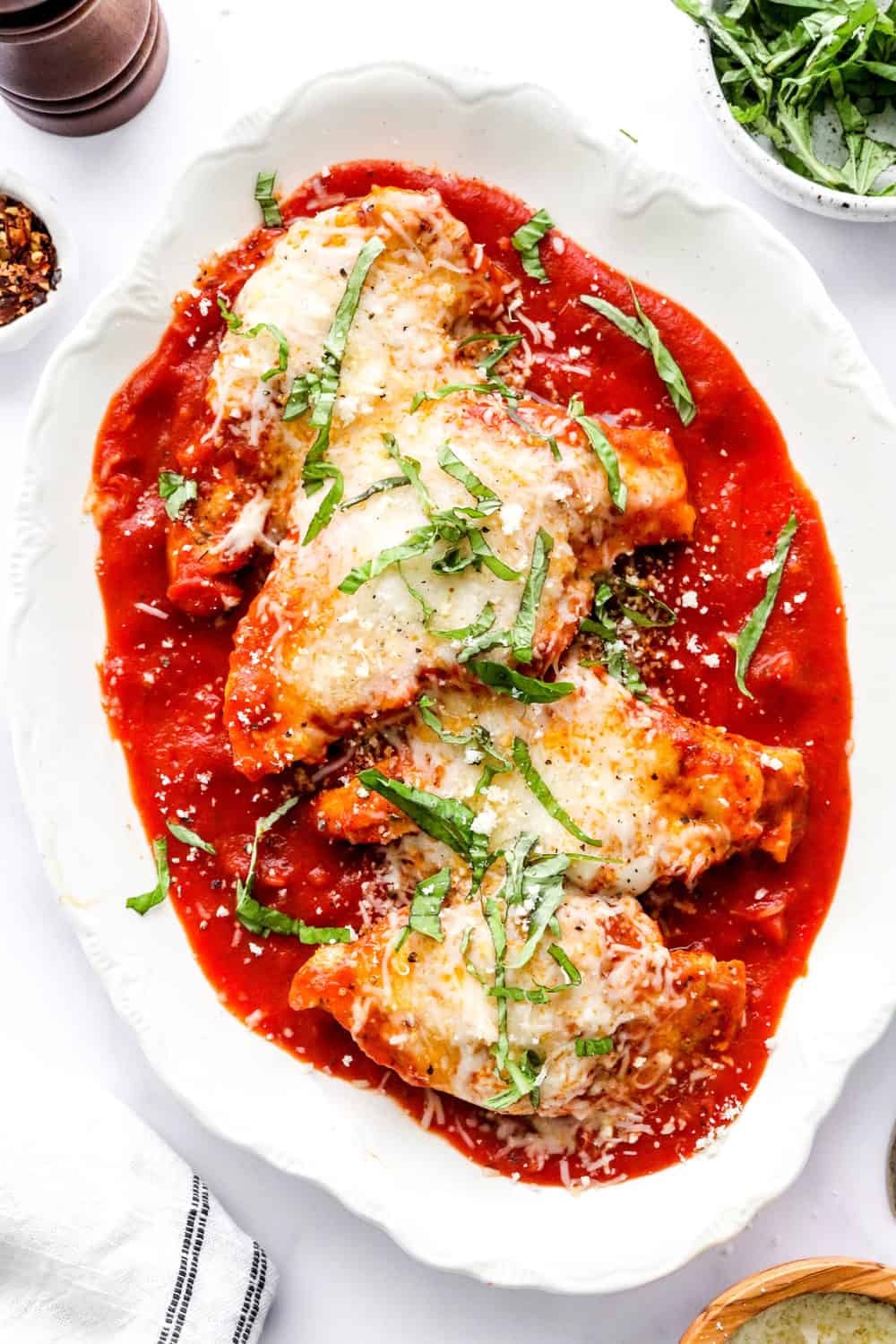 Oval, white platter with tomato sauce spread on the bottom topped with hearty chicken topped with more red sauce and melted cheese, topped with sliced fresh basil