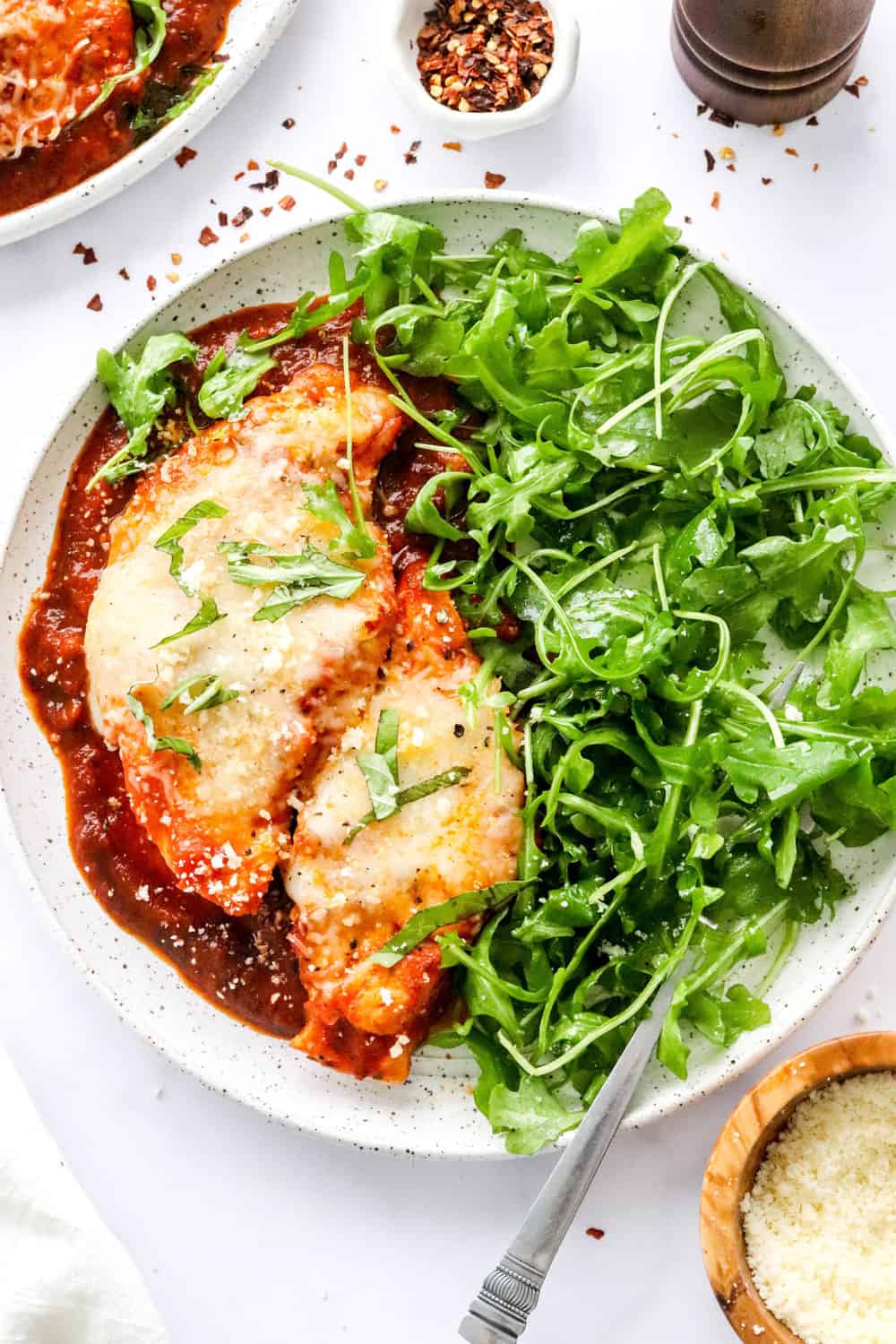 Instant pot chicken parmesan on a plate on top of tomato sauce with seasoned arugula on the plate next to it with another plate of of chicken parmesan behind I and a wooden bowl filled with parmesan cheese in front of it. 