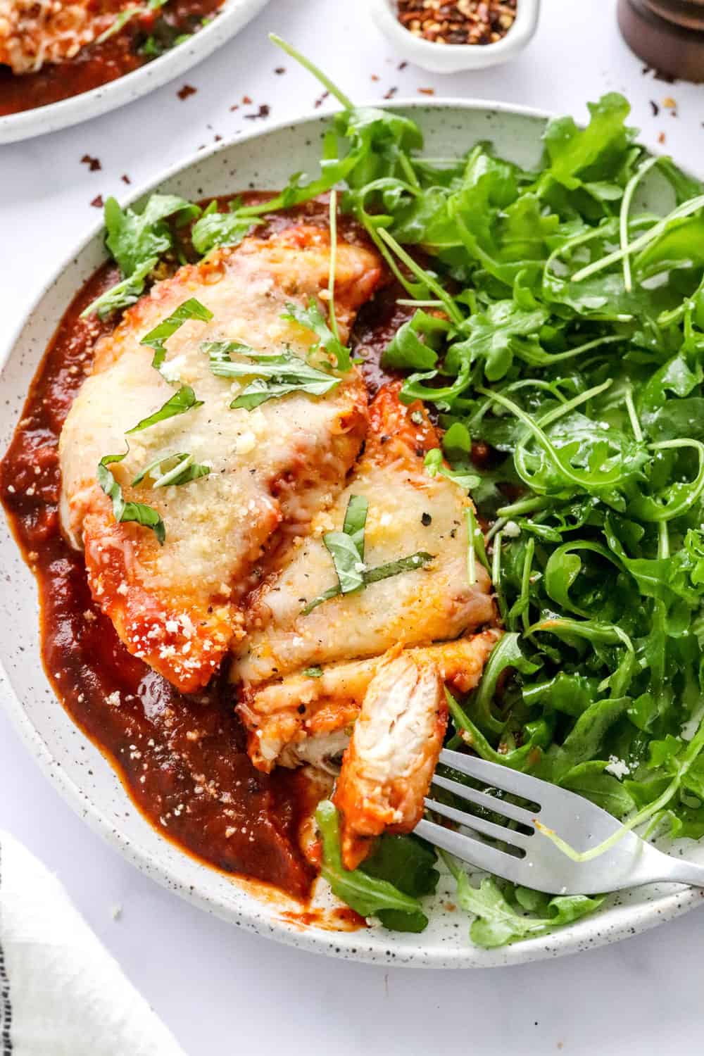 Chicken cooked in tomato sauce topped with melted cheese on a plate next to an arugula salad with a form on the plate