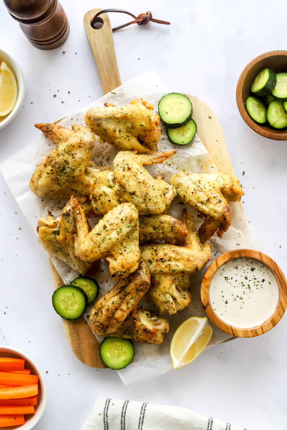 A pile of baked chicken wings seasoned with lemon pepper seasoning on white parchment paper on top of a wooden cutting board with a white dipping sauce in a bowl on the board and bowls of veggies around it. 