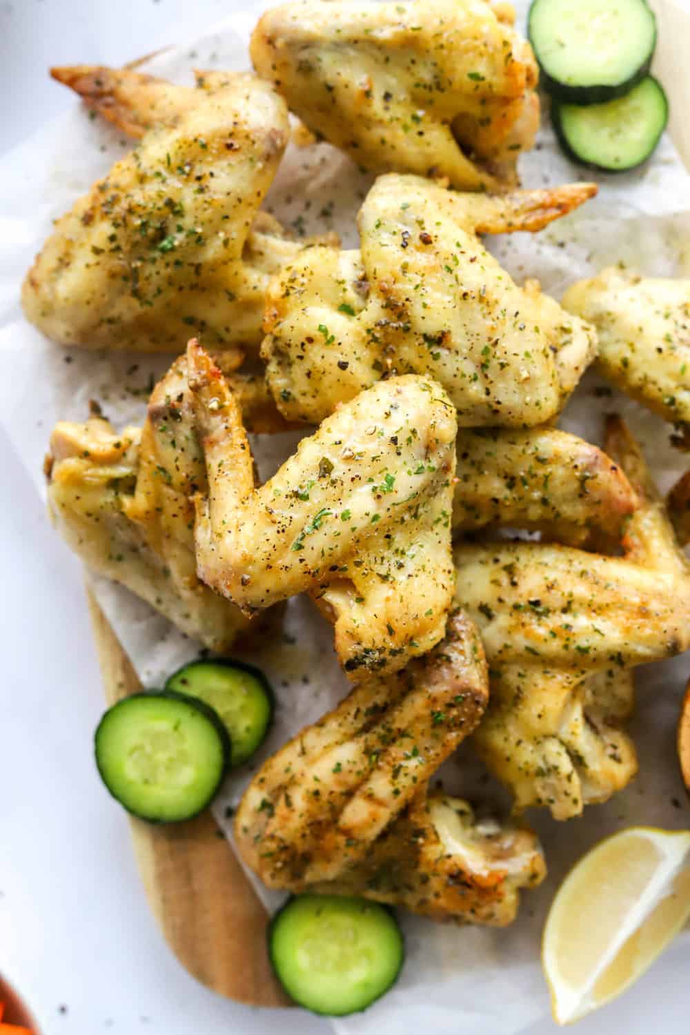 Crispy lemon chicken wings piled up on a wooden platter with cucumber slices and a lemon wedge around them. 