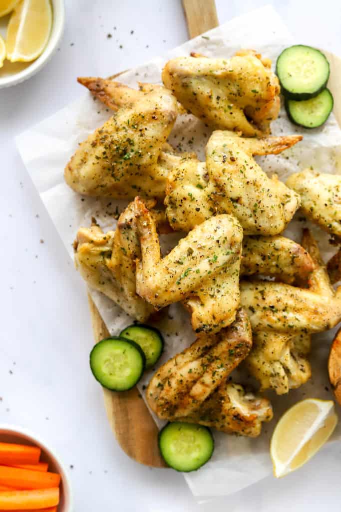 Close up of a pile of golden brown, seasoned chicken wings on a serving board on top of parchment paper with a few cucumber slices next to them. 