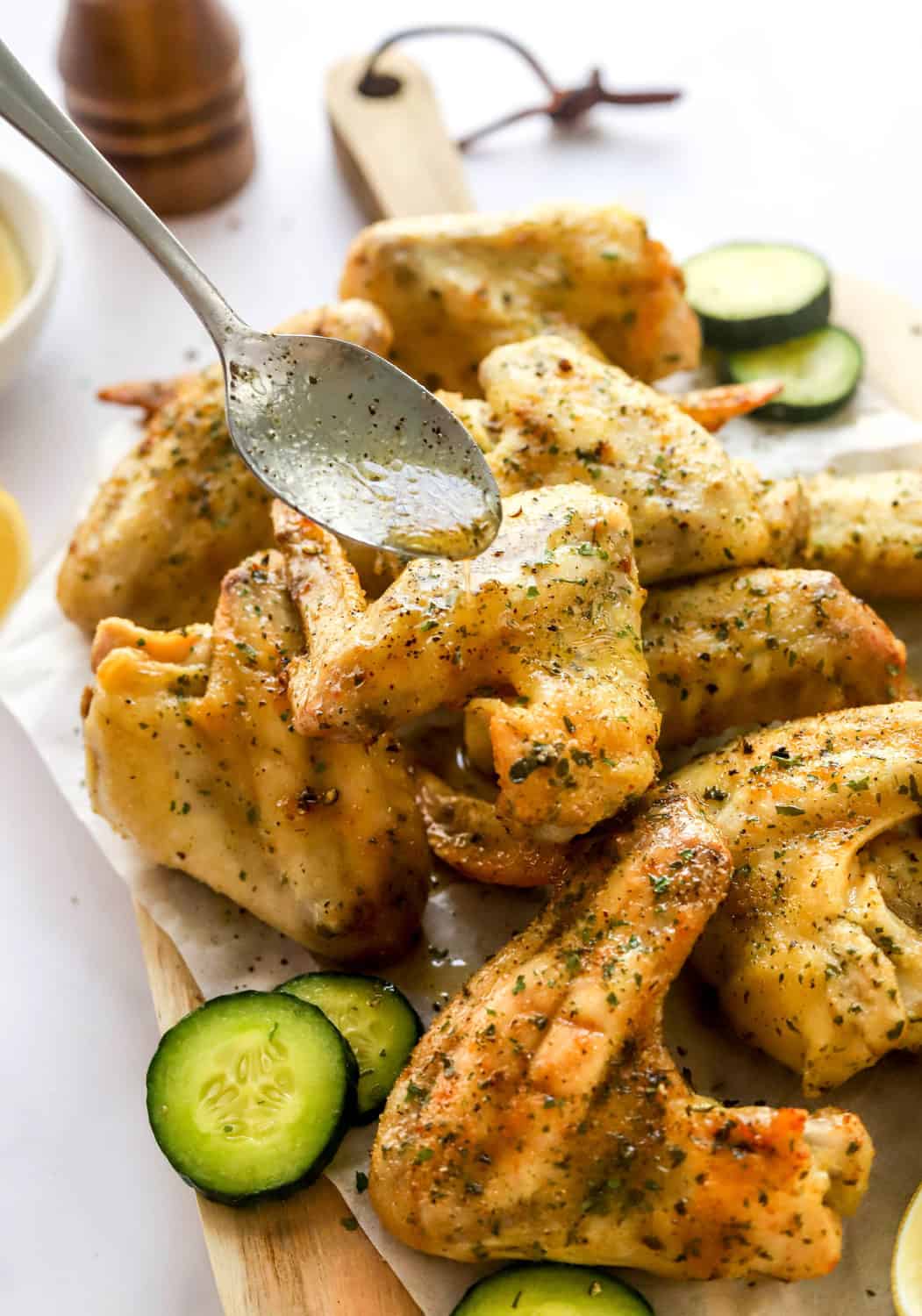 Lemon pepper chicken wings piled on a wooden board with cucumber slices in from of them and a spoon dripping lemon pepper sauce on them. 