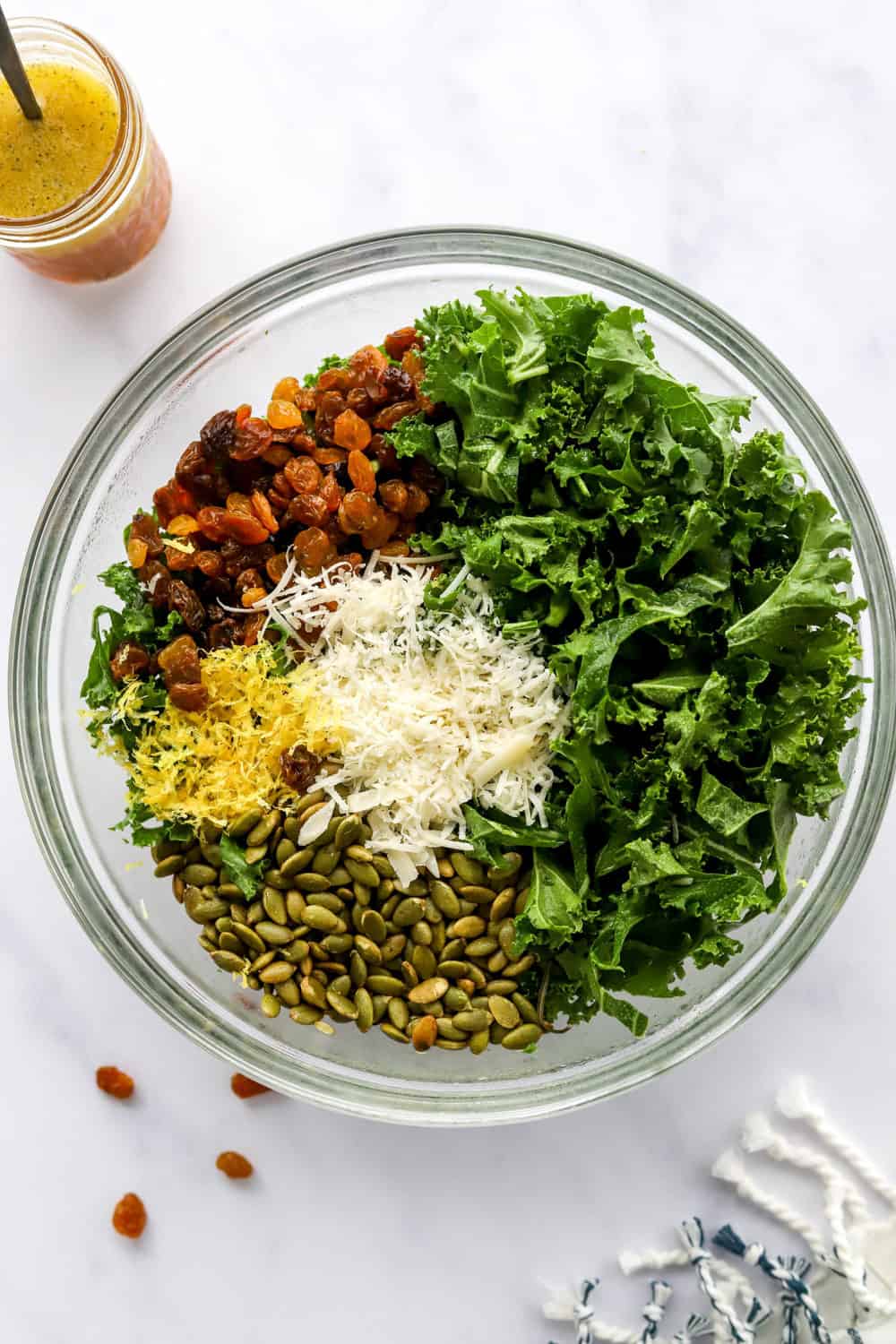 Round serving bowl with green kale, pumpkin seeds, lemon zest, shredded parmesan cheese and golden raisins in it with salad dressing in a jar behind it