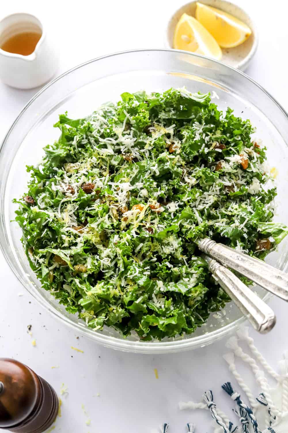 Chopped kale in a glass bowl mixed up with nuts, raisins, shredded cheese and lemon zest with silver serving spoons in the bowl and salad dressing in a white jar with lemon slices in a bowl behind it