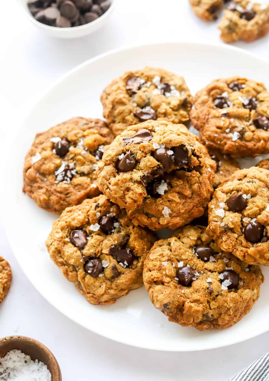 A plate full of healthy oatmeal chocolate chip cookies with a bowl of chocolate chips behind it. 