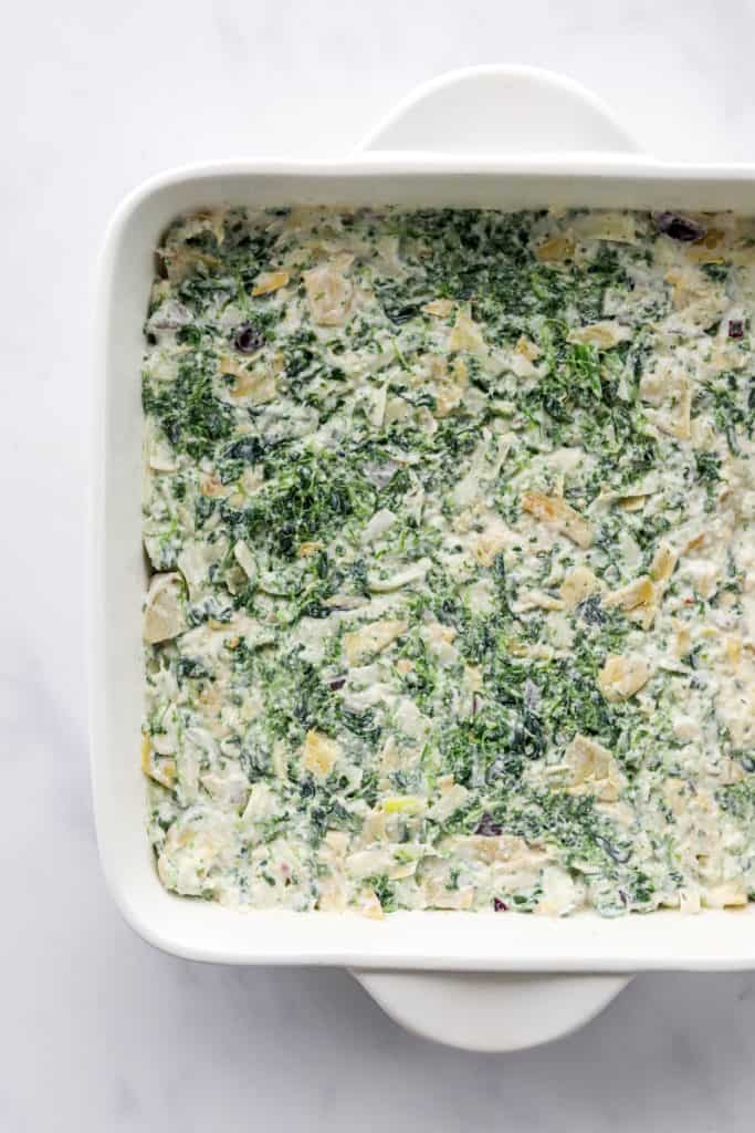 Spinach and artichoke mixture spread out in a white, square baking dish