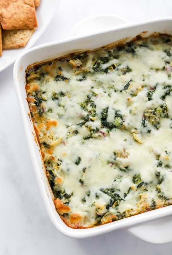 White square baking dish filled with cheesy spinach dip with pita chips on a plate behind it