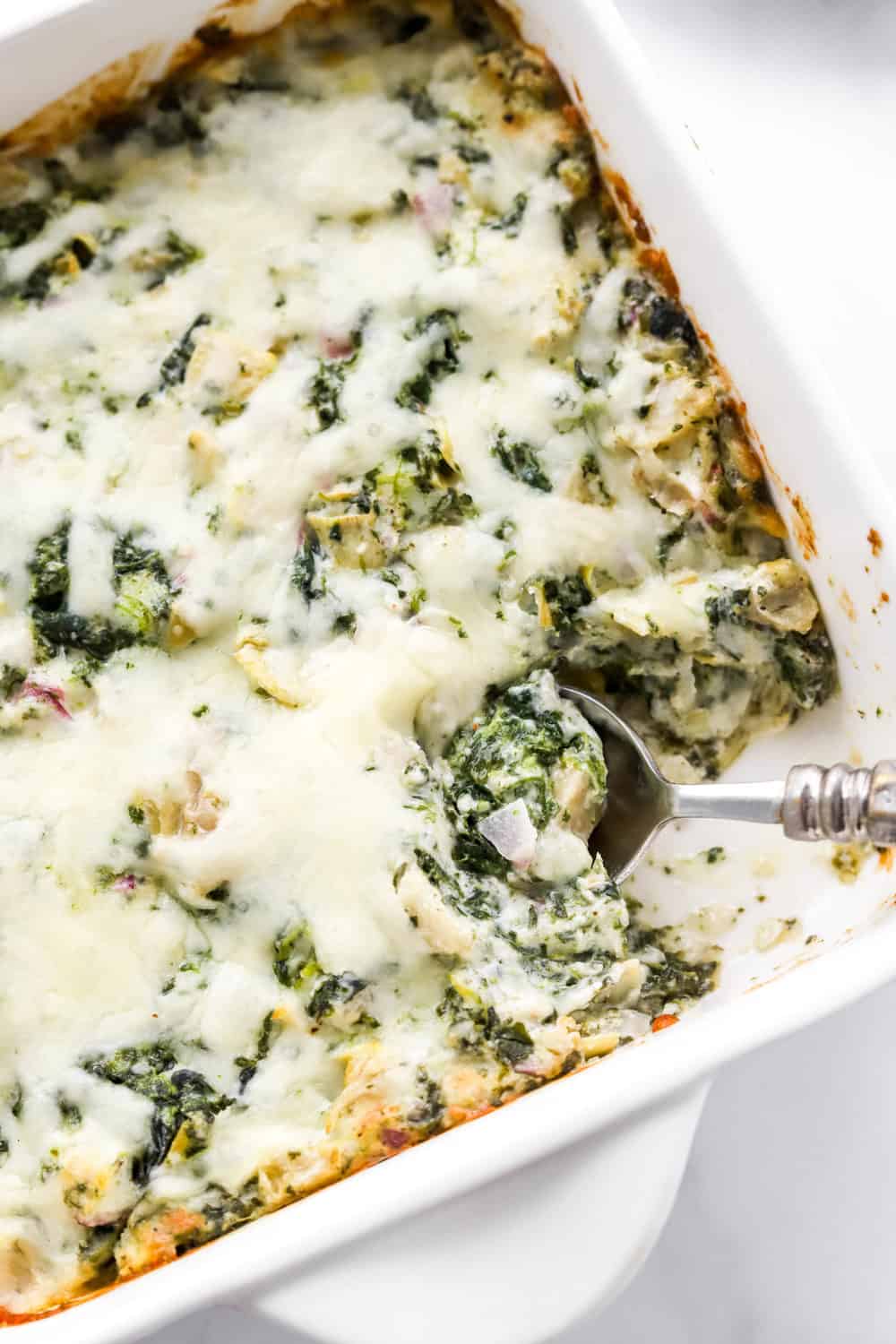 Silver spoon scooping cheesy greens out of a baking dish finked with cheesy, green dip